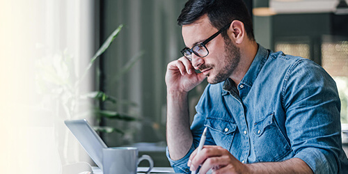 A person in a denim shirt sits at a desk, focused on a laptop, writing notes in a notebook with a mug nearby, surrounded by greenery