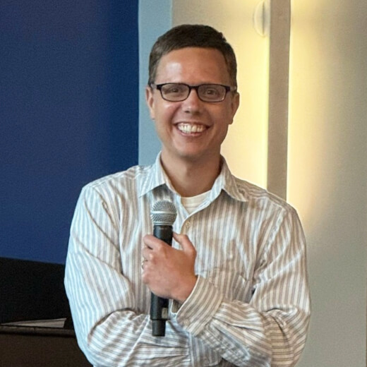 Smiling man wearing glasses and a striped shirt holds a microphone while speaking indoors.