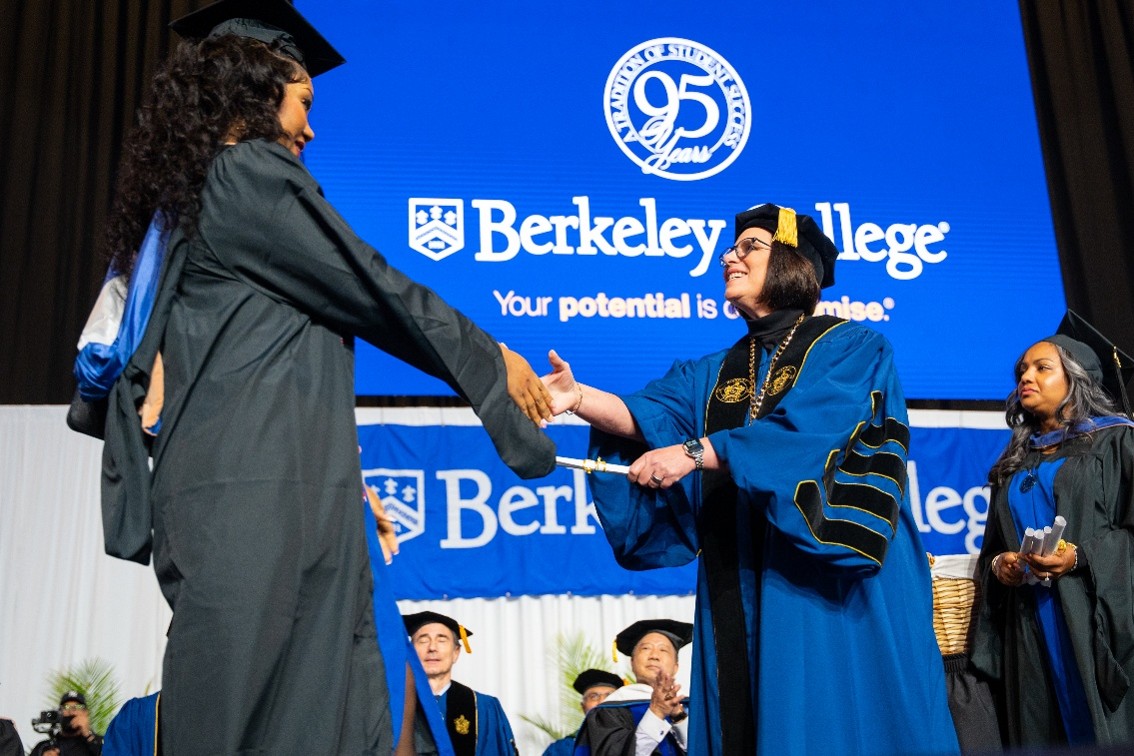 A Black woman in Commencement regalia receives her diploma and shakes hands with a female college president, dressed in doctoral robes.
