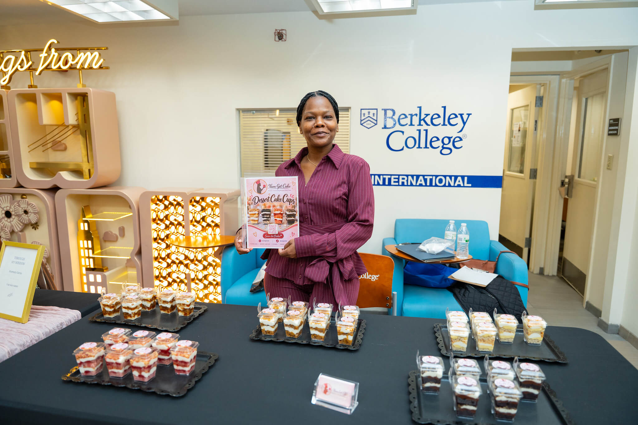 Woman displays a dessert cake cup menu at a Berkeley College event, with trays of layered desserts on the table.