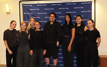 Group of eight diverse students standing and smiling in front of a Berkeley College backdrop indoors.