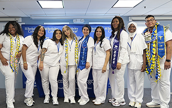 A group of diverse students in white scrubs stands in a line and smiles for the camera.