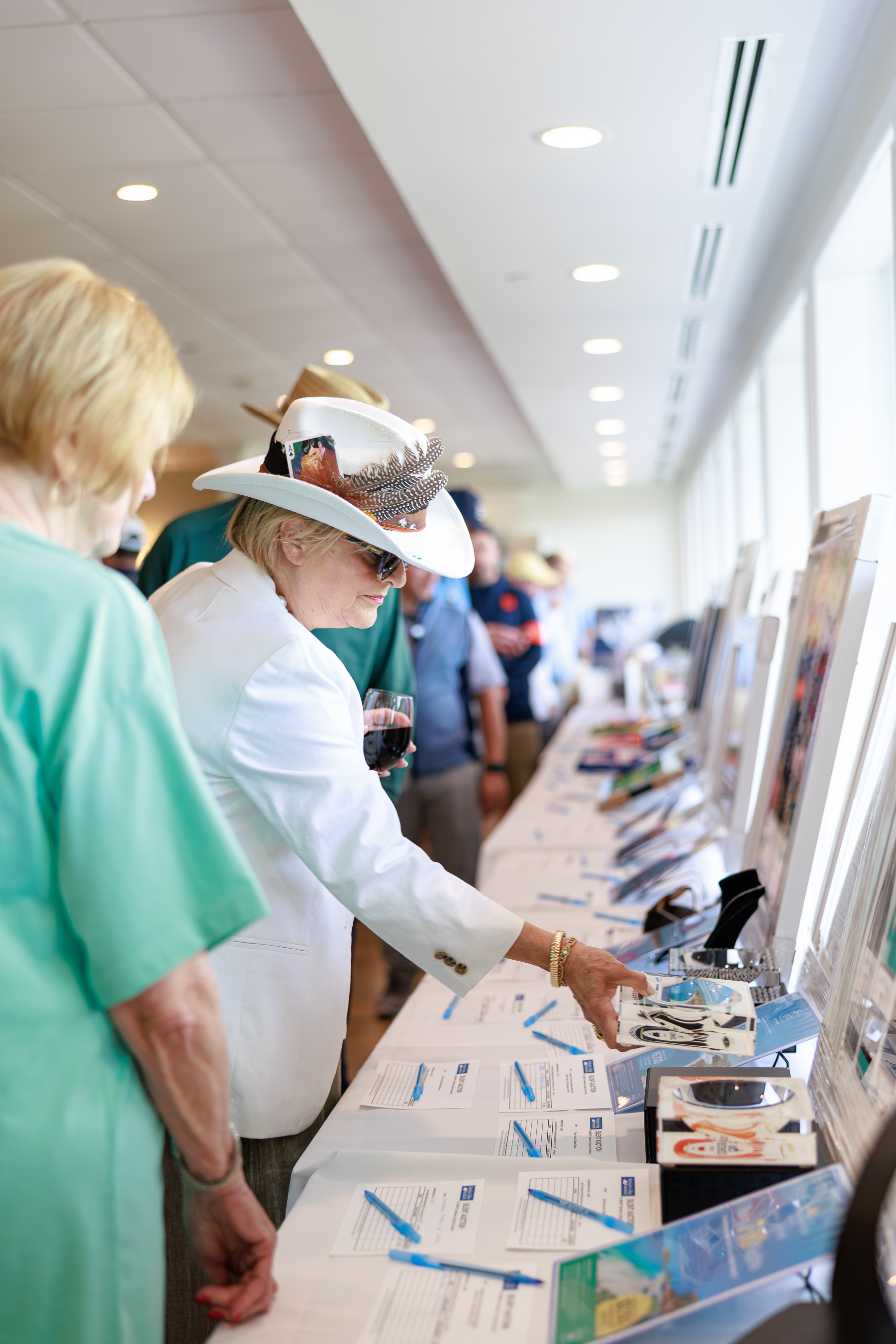 A woman in a white hat stands over an auction table inspecting art.