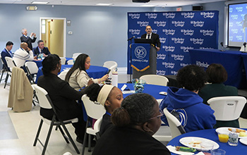 A speaker at podium addressing a seated audience in a conference room with Berkeley College banners.
