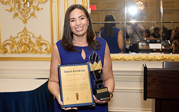 Woman in blue dress holding framed award certificate and trophy in an ornate room with gold decorations and a mirror behind.