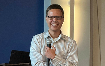 Smiling man wearing glasses and a striped shirt holds a microphone while speaking indoors.