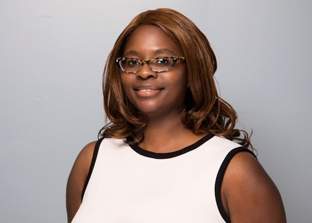 Portrait of a smiling woman with brown hair, glasses, and a sleeveless white top against a plain gray background.