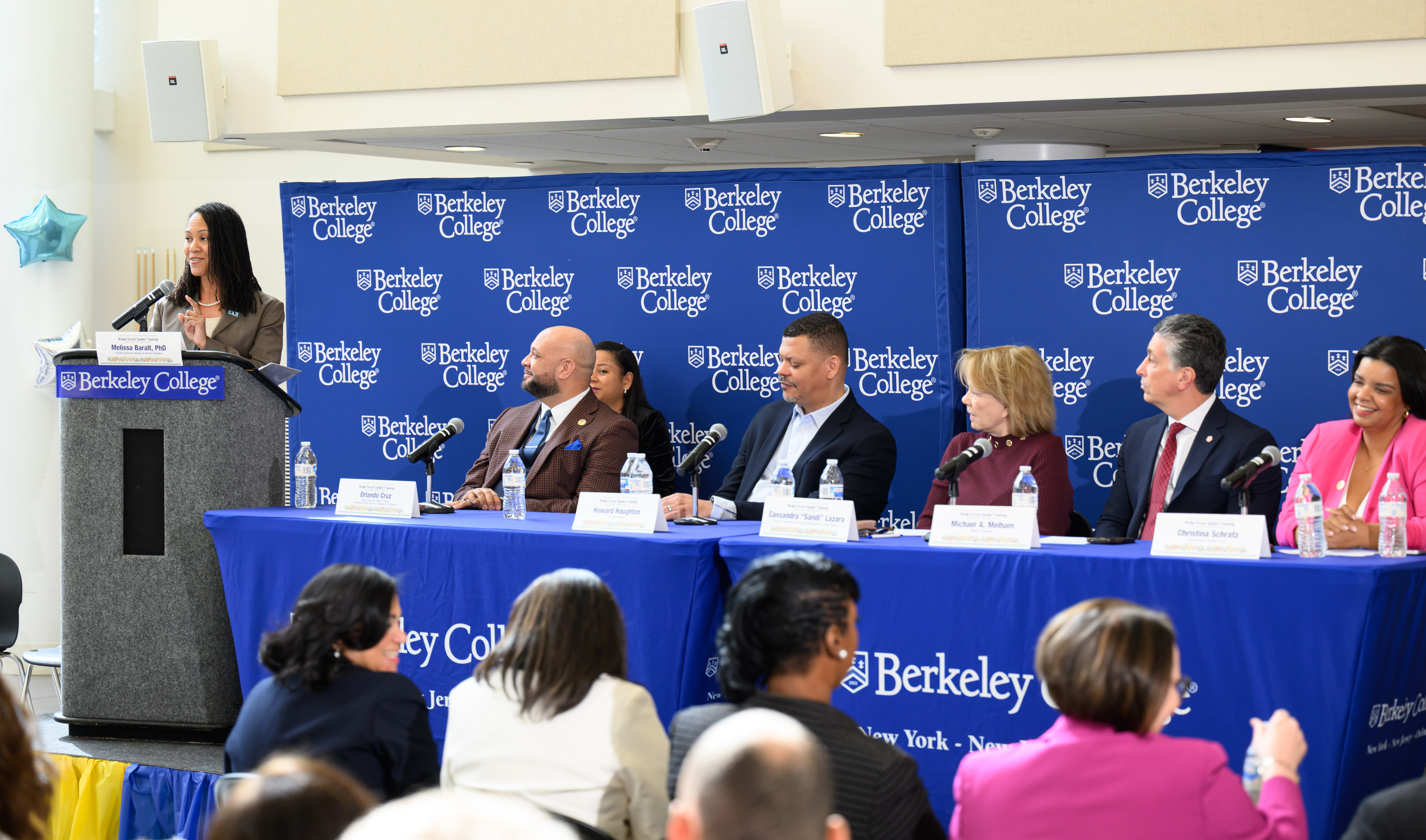 Speakers stand at a podium and sit at a table in front of a blue backdrop