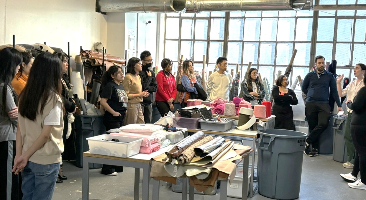 Students stand in a warehouse with a table in the center of the room covered in different rolled fabrics.