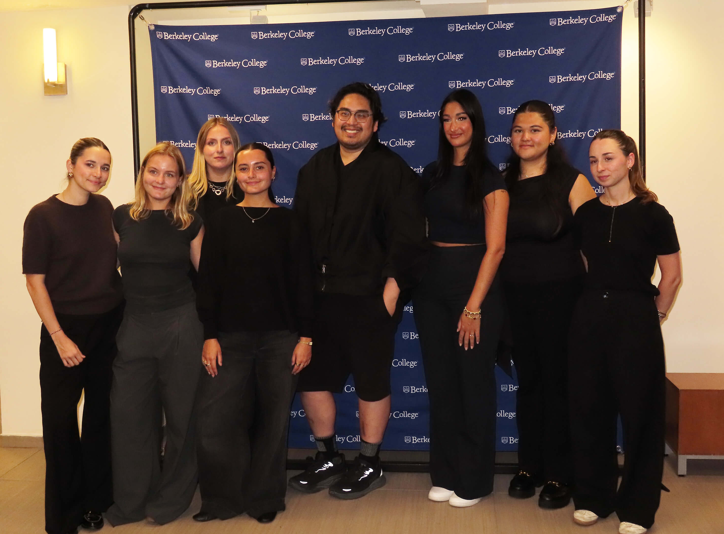 Group of eight diverse students standing and smiling in front of a Berkeley College backdrop indoors.