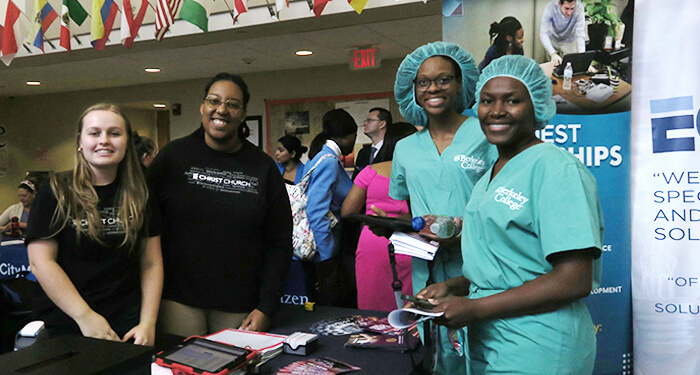 our women stand beside a table displaying a sign. Two women wear nursing scrubs showcasing a professional healthcare environment, as they talk to the other two women in casual clothes, who promote their company as a potential employer.