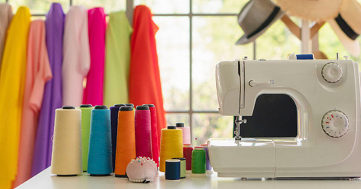 A sewing machine sits on a table with colorful spools of thread, surrounded by vibrant fabric draping in the background.