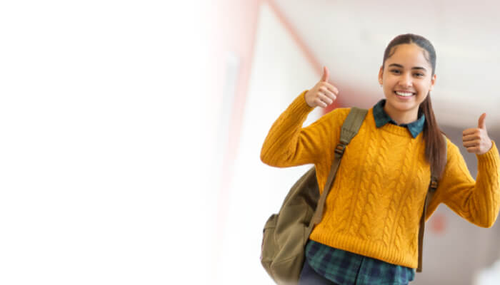 A person in a yellow sweater gives a thumbs-up while wearing a backpack in a brightly lit hallway