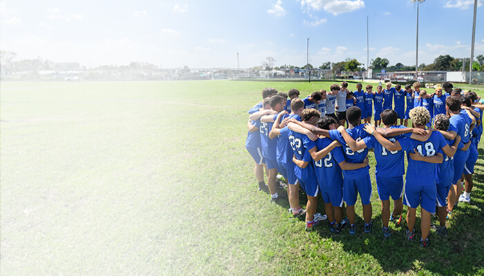 A soccer team in blue uniforms huddles together on a sunny field, raising their hands in unity. Trees and a building are visible in the background.