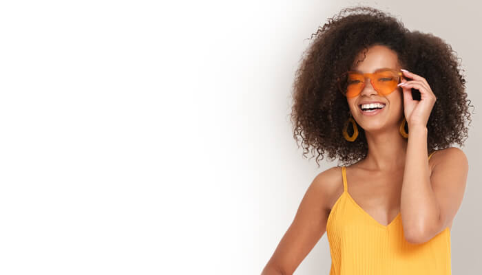  A woman wearing a yellow dress and orange sunglasses stands confidently, showcasing her vibrant summer style