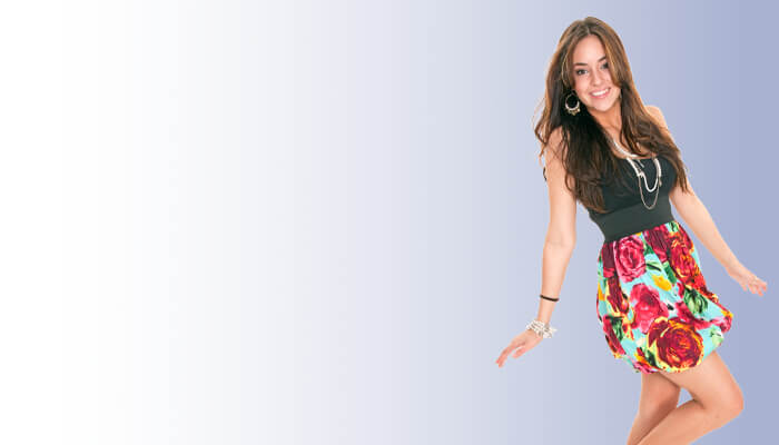 Smiling woman in a colorful floral skirt and black top standing against a gradient white to blue background, exuding a joyful, playful energy.