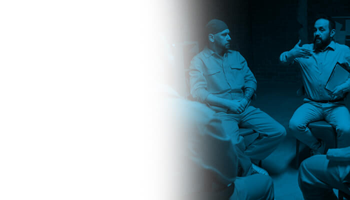 A group of individuals in uniform sit in a circle, engaged in conversation in a dimly lit room with a blue hue.