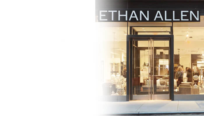 Ethan Allen furniture showroom storefront with glass entrance, warm interior lighting, furnishings, and shoppers visible inside; polished, inviting tone