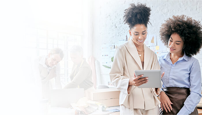 Two women stand smiling while looking at a tablet, with a bright office setting in the background. Two other people are working at a desk behind them