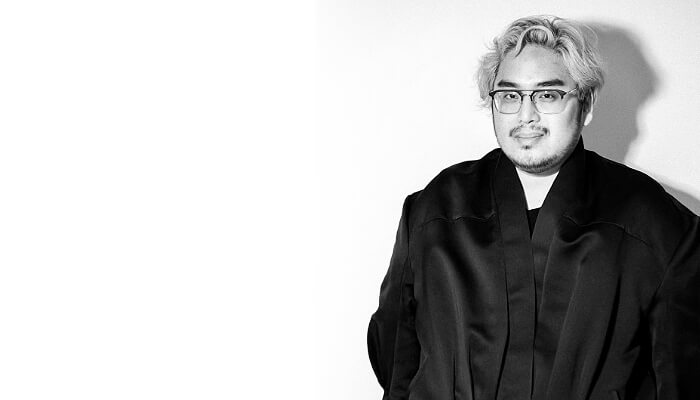 Black and white photo of a person with light hair, glasses, and a black coat, smiling slightly. They stand against a plain, light background.