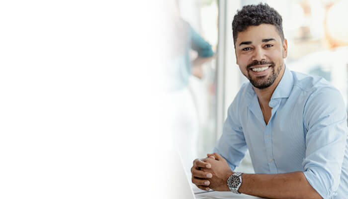 A smiling young businessman in a light blue dress shirt sitting at a desk, with a white gradient fade on the left side