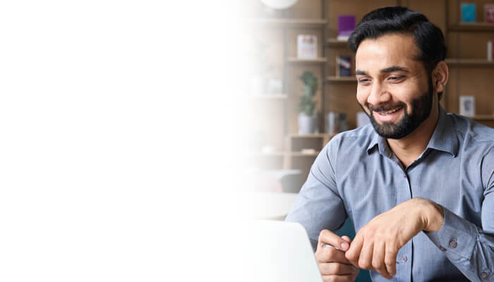 A man with a beard, wearing a gray shirt, sits at a desk and smiles while looking at a laptop in a modern indoor setting