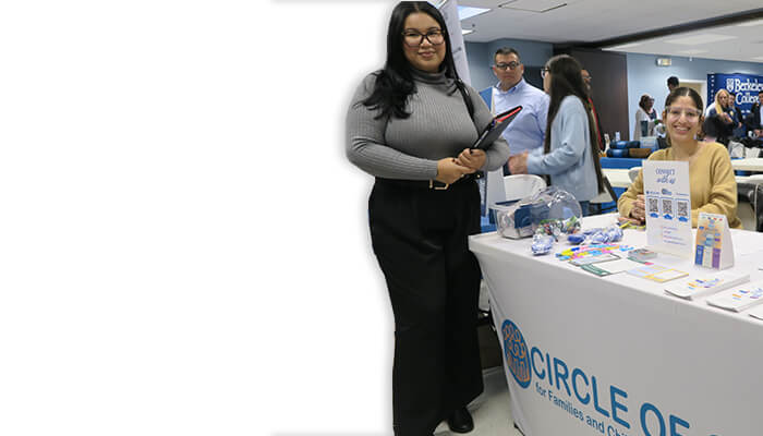 Smiling woman in a gray sweater stands beside a Circle of Care booth, where volunteers share health resources at a community event