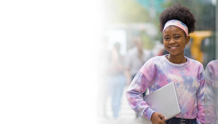Smiling Black teenage girl in a pink tie-dye sweatshirt and headband carries a closed laptop outdoors, conveying confidence and optimism