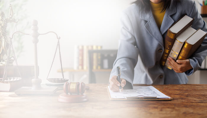 Lawyer in a gray suit writes at a wooden desk while holding law books; scales of justice and a gavel convey a serious legal setting