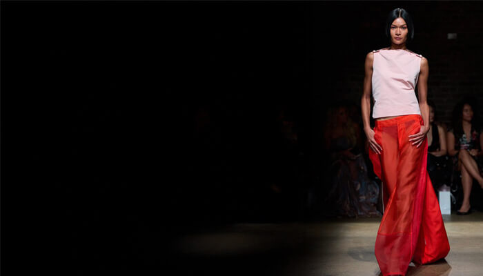 A model walks the runway in a stylish pink top and flowing red skirt, showcasing contemporary fashion in a dimly lit venue