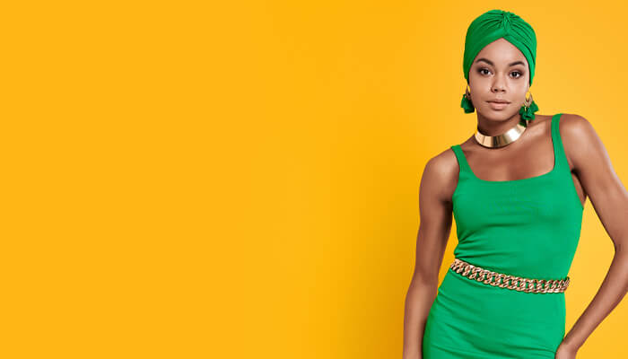 Woman in green dress and turban stands confidently against yellow background. She's accessorized with gold earrings, choker, belt, conveying elegance.