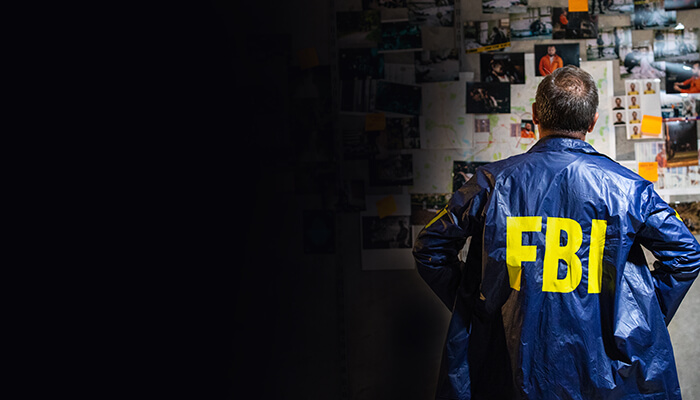 FBI agent in a blue jacket stands in front of a wall covered in photos and notes, deep in thought.