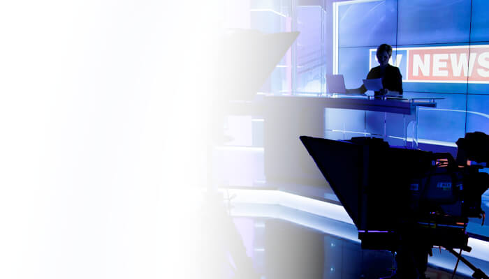 Silhouetted news anchor at a modern TV studio desk, bright blue background with News text, camera equipment in foreground; professional atmosphere.