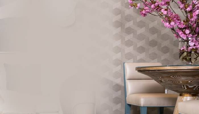 A stylish interior featuring a round wooden table, a beige chair with blue accents, and a vase of pink flowers against a geometric wall.
