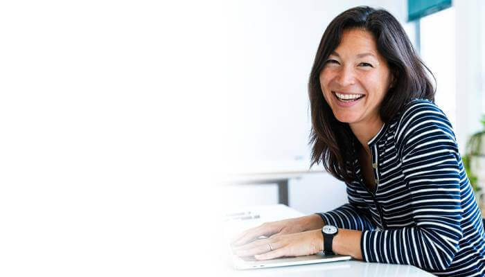 Smiling woman with long hair in a striped shirt sits at a desk, hands resting on a laptop, conveying a cheerful and approachable demeanor