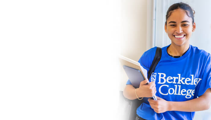 Smiling woman in a blue "Berkeley College" shirt holds books and a backpack strap. She's standing indoors against a light background, conveying a positive academic vibe.