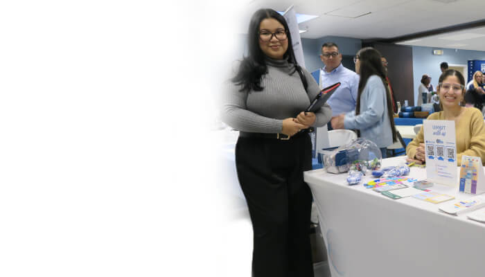 Attendees networking at the Berkeley College NYC Winter Career Fair booth. A woman holding a folder smiles beside a booth at an event.