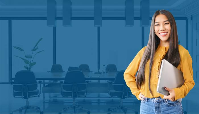 A smiling woman in a yellow blouse holds a laptop, standing confidently in a modern office setting with a conference table and chairs.