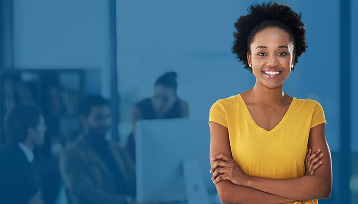 Smiling woman in a yellow shirt stands confidently with arms crossed. Office background with blurred colleagues working at computers conveys a professional tone