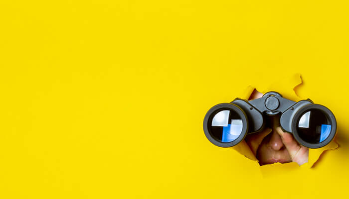A figure is seen peering through a circular hole in a bright yellow wall, suggesting curiosity and intrigue