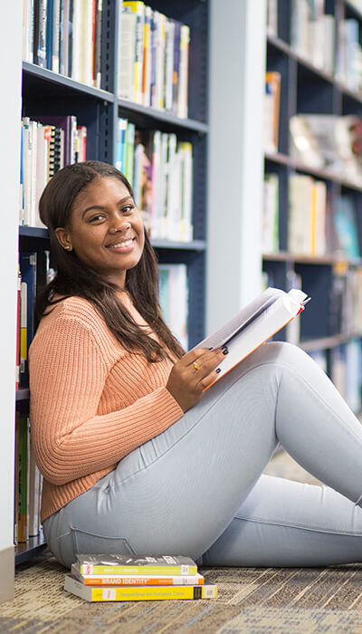 A young woman sits on the floor, surrounded by bookshelves filled with various books, immersed in reading