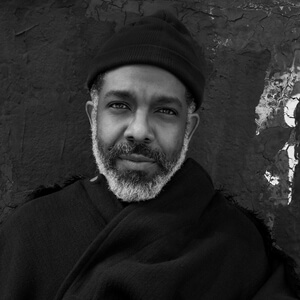 Black-and-white portrait of Aaron Potts in a knit cap and dark scarf, gazing calmly at the camera against a textured wall.