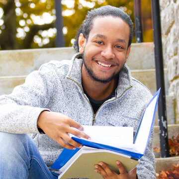 Photo of a male student holding a book while sitting down on campus steps mobile image mobile image