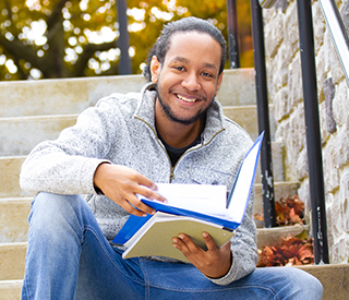 Photo of a male student holding a book while sitting down on campus steps mobile image