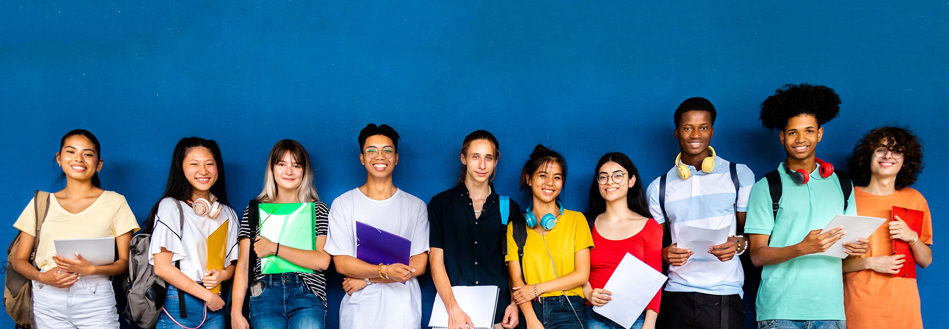 Image: A diverse group of nine smiling students stands against a blue wall holding notebooks