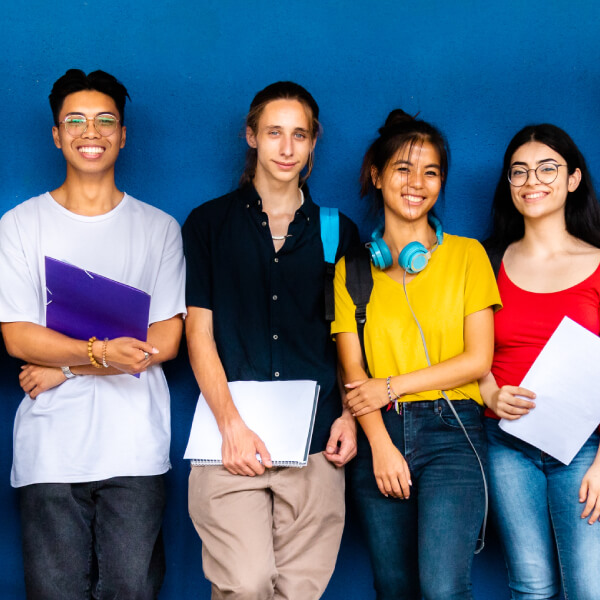 Image for mobile devices: A diverse group of nine smiling students stands against a blue wall holding notebooks