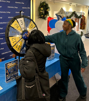 Two people stand at a spin-the-wheel game at a Berkeley College event. One spins the wheel, while the other watches. It's a lively, festive atmosphere.