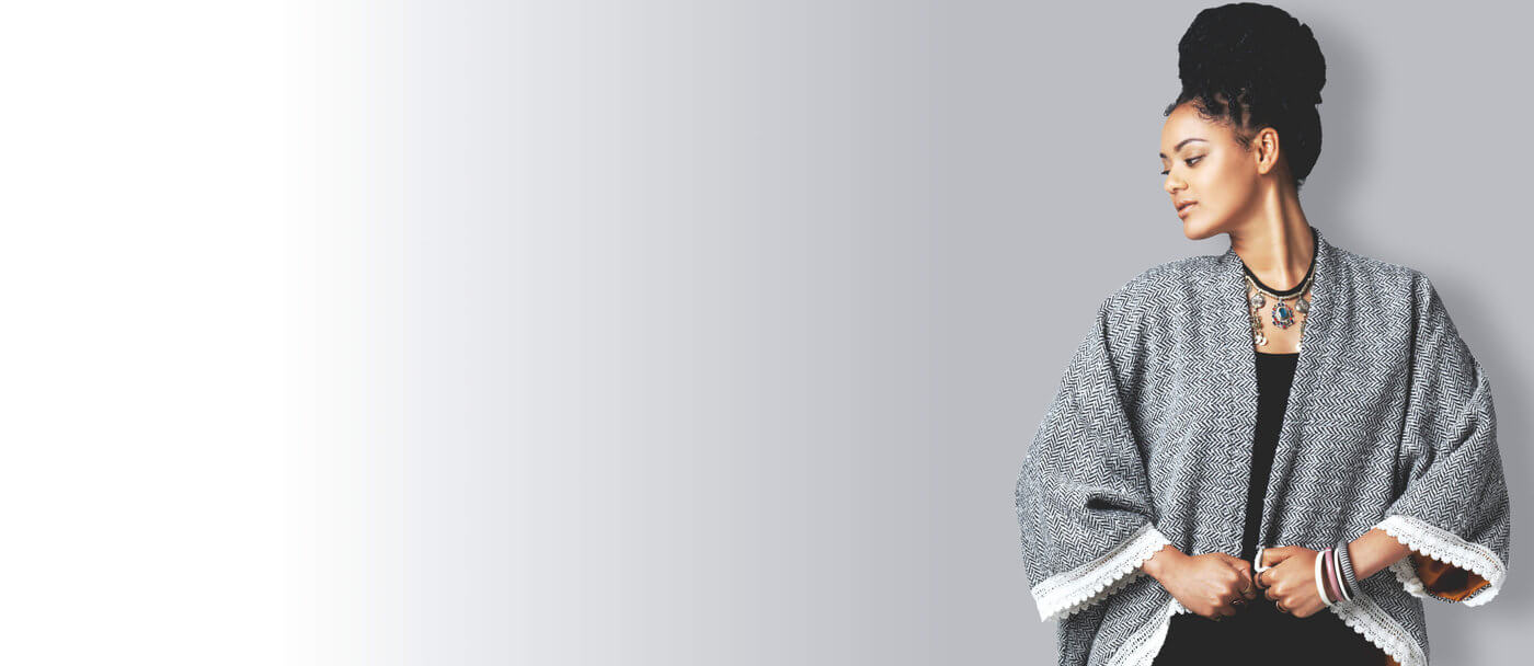 Woman in a black dress and gray shawl stands against a gray gradient background. Her expression is calm and confident, with a stylish, modern look.