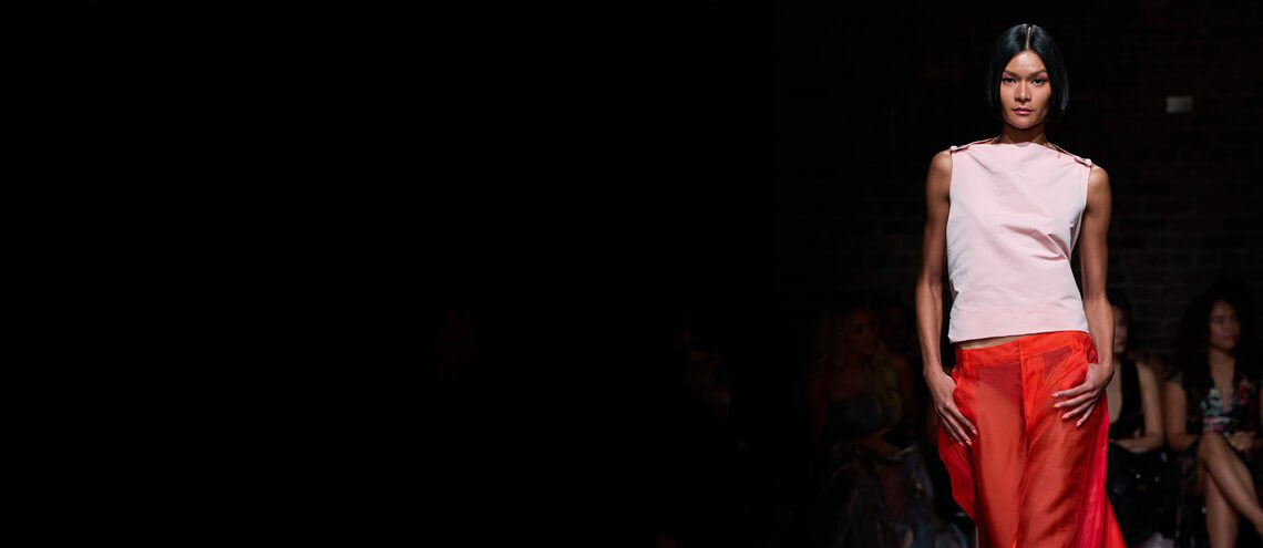 A model walks the runway in a stylish pink top and flowing red skirt, showcasing contemporary fashion in a dimly lit venue