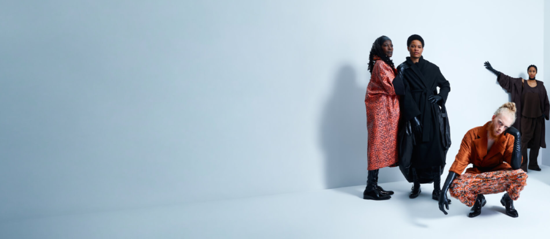 Four fashion models pose against a pale blue studio backdrop: two stand in bold coats, one crouches in orange, and one gestures behind them, creating an editorial mood